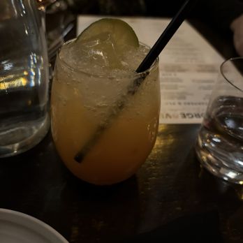 Forge & Vine - Apple cider mocktail mule - A drink served in a rounded glass contains a light-colored liquid with ice, garnished with a lime wedge and a black straw. The setting appears to be a restaurant or bar with a dark wooden table, a menu in the background, and other glassware nearby. The drink is labeled as an apple cider mocktail mule, consistent with the restaurant's offerings. - American Near Me - Groton, Massachusetts