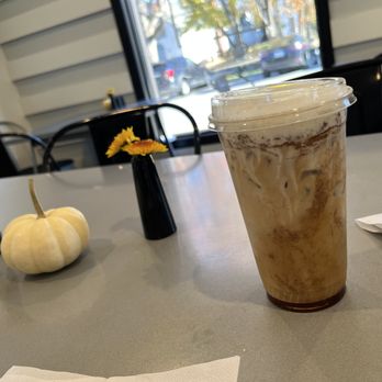Twisted Tree Cafe - An iced coffee beverage in a clear plastic cup with a lid, placed on a table inside a cafe. The table has small vases with yellow flowers and a white pumpkin, with a window showing parked cars outside. - Coffee & Tea Near Me - Lincoln, Massachusetts