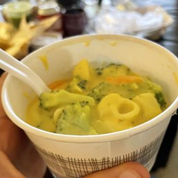 A paper cup filled with a creamy vegetable soup containing broccoli, carrots, and potatoes, with a plastic spoon inside.