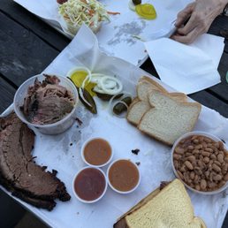 Brisket Sandwich - A barbecue meal includes sliced beef, pulled pork, bread slices, baked beans, and pickles served on a tray with sauces.