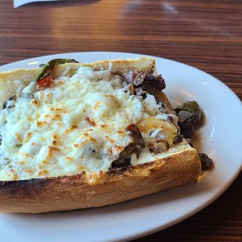Dengeos - Philly Steak Sandwich - A baked potato is topped with melted cheese, grilled vegetables such as zucchini and onions, and possibly pieces of meat. The dish is served on a white oval plate placed on a wooden table surface, with some paper or napkin partially visible at the top of the image. - Greek Near Me - Buffalo Grove, Illinois