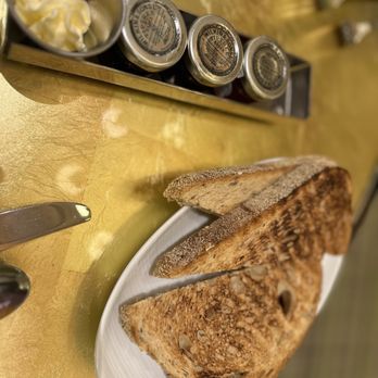 Giardino - Toast - Slices of toasted bread with seeds are arranged on a white plate, accompanied by butter and jam jars on a yellow table. - Juice Bars & Smoothies Near Me - Everett, Massachusetts