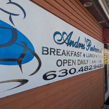 Andres Restaurant - Welcome - The image features a large signboard mounted on the exterior wall of Andres Restaurant. The sign displays the restaurant's name, hours of operation from 8 a.m. to 4 p.m., contact phone number, and mentions breakfast, lunch, and dinner services. The sign has a graphic of a fish and is attached to a building with a wooden exterior and a small outdoor seating area with an awning. - Spanish Near Me - Batavia, Illinois