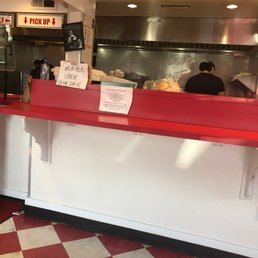 Inside a restaurant, a counter with a red top and white base is shown, with kitchen staff working behind it and signs indicating pickup and other instructions.
