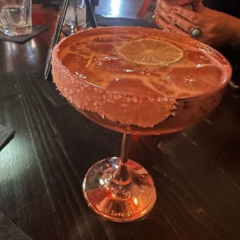 The Overpour - A cocktail glass with a salted rim holds a drink garnished with lemon slices and floating citrus slices. The glass is placed on a dark wooden table in a dimly lit indoor setting, with other glasses and a person holding a small bowl visible in the background. The setting appears to be a bar or restaurant environment. - Cigar Bars Near Me - El Paso, Texas