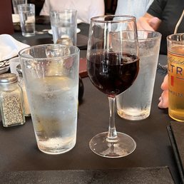 three glasses of wine on a table - A table in a restaurant features glasses of water, a glass of red wine, and a glass of beer, with some condiments and utensils visible. Several people are seated around the table, with their upper bodies and arms visible, engaged in dining or conversation. The background shows a dimly lit interior with wooden furniture.