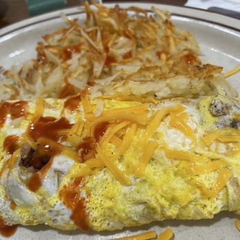 Sunshine Diner - Omelet - A breakfast plate features a large omelet topped with shredded cheddar cheese and drizzled with sauce. Next to the omelet, there are hash browns with a crispy texture. The plate is set on a wooden table with some condiments and napkins visible in the background. - Diners Near Me - Lansing, Michigan