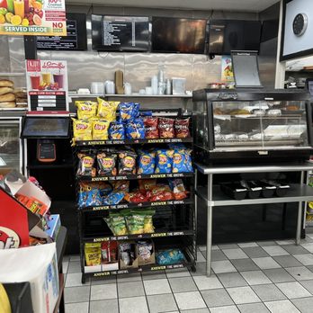 Quick N Out - the inside of a fast food restaurant - The image shows a retail space with a snack display rack holding multiple bags of chips, including Lay's, Doritos, Cheetos, and others. To the right, there is a glass display case with baked goods, possibly muffins or pastries. Behind the display, there are digital screens and a counter with cups and utensils, indicating a food service area. The floor is tiled, and shelves with bananas and other products are visible on the right side. - Delis Near Me - Union, New Jersey
