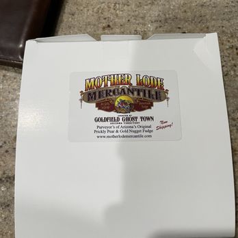 Mother Lode Mercantile - Chocolate fudge packaging! - A white cardboard box with a label from Mother Lode Mercantile, located at Goldfield Ghost Town in Arizona, is shown on a textured surface. The label indicates the store offers homemade fudge, including Prickly Pear and Gold Nugget flavors, and features the store's website. A small note on the label mentions 'Now Shipping!'. A brown wallet is partially visible in the background. - Candy Stores Near Me - Apache Junction, Arizona