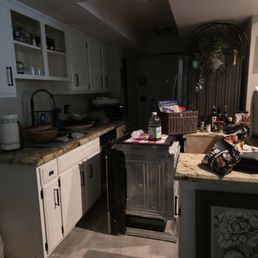 What floor daddy did to my home - A kitchen features white upper and lower cabinets with black handles, granite countertops, and a stainless steel sink with a black faucet. The counter holds a water bottle, snacks, and small appliances. There are open shelves with glassware and jars. The scene is dimly lit, with a dark background and various cluttered items on the counter, including a basket, bottles, and paper towels. The floor is visible, and part of a decorative framed picture is seen on the wall.
