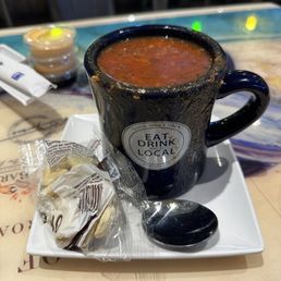 Mama's Conch Chowder - A black ceramic mug filled with a dark, broth-based soup is placed on a white square tray. The mug has a sticker that reads "Eat, Drink, and Local" with the phrase "Good Vibes Only." A metal spoon rests on the tray, and a wrapped snack is positioned nearby. In the background, there are small containers and a colorful table surface, suggesting a casual dining setting.