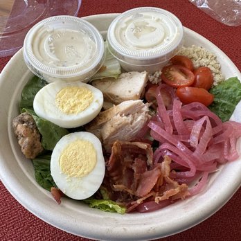 Jason's Deli - The BLT bowl, no avocado, with extra ranch. - A disposable plate holds a salad with sliced boiled eggs, cherry tomatoes, shredded chicken, bacon strips, pickled red onions, and lettuce leaves. Two small plastic containers of dressing are placed on the plate. The plate is on a red tablecloth, with a plastic cover and glassware nearby. - Delis Near Me - Albuquerque, New Mexico