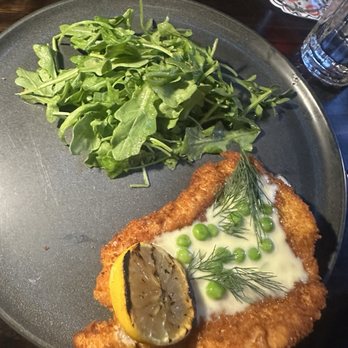 Sudova * Eastern European Kitchen - kotleta (fried chicken + dill sauce) - A breaded fish fillet topped with dill, green peas, and a lemon slice, served on a dark plate with a side of arugula salad. - Modern European Near Me - Cincinnati, Ohio