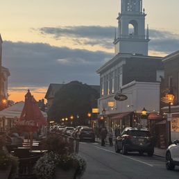 A street scene during sunset in a small town, with a church steeple visible in the background. The street has parked cars, pedestrians walking, and storefronts with illuminated signs. Outdoor seating with umbrellas is present, and potted plants decorate the sidewalk. The sky is partly cloudy with a warm glow from the setting sun.
