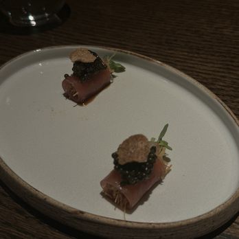 Valhalla - Two small appetizer portions are arranged on a white, oval-shaped plate placed on a wooden table. Each consists of a rolled cured meat, topped with black caviar, a small garnish, and a tiny crispy element. The setting appears to be in a dimly lit dining environment, possibly a fine dining restaurant. - Bars Near Me - Chicago, Illinois