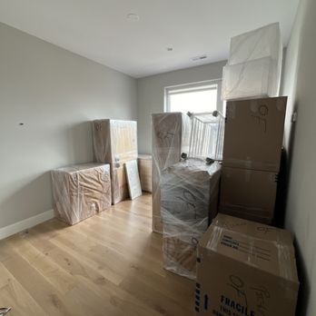 Bernard Movers - A room contains multiple packed boxes and furniture wrapped in plastic, with a window providing natural light, indicating a moving or storage process. - Movers Near Me - Chicago, Illinois