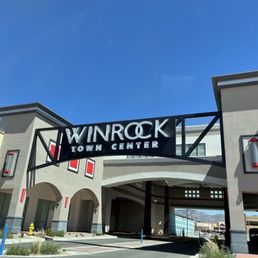 The photo captures the main entrance of Winrock Town Center, a shopping and lifestyle destination in Albuquerque, NM. The sign is mounted on a modern building facade with neutral tones, featuring decorative lighting fixtures on either side. The sky is clear and blue, and the surrounding area includes some landscaping and distant mountains. The image emphasizes the commercial and community-oriented nature of the town center.