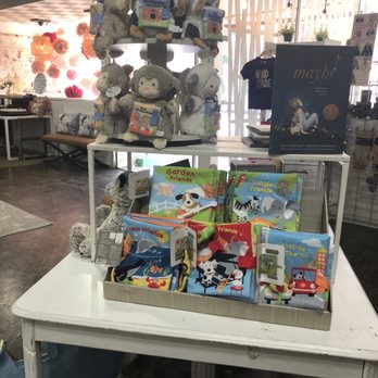 HAND+made - Kids toys - The image shows a retail store interior with plush animal toys, including bears, sheep, and other animals, arranged on a multi-tiered stand. In front of the toys, a white table displays children's books with colorful covers about animals and friends, such as 'Garden Friends' and 'Jungle Friends.' The background includes a seating area with a bench and decorative wall elements, with natural light coming through large windows. - Gift Shops Near Me - Laurel, Mississippi