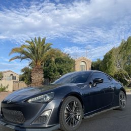 Windshield: 50% Side and back: 35% - A black Toyota sports car is parked on a paved street in a suburban neighborhood. The car has tinted windows and alloy wheels. Behind the car, there is a tall palm tree, green trees, and residential houses with beige and brown exteriors. The sky above is partly cloudy with patches of blue.