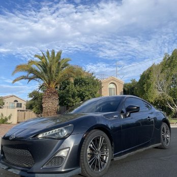 Phantom Tint - Windshield: 50% Side and back: 35% - A black Toyota sports car is parked on a paved street in a suburban neighborhood. The car has tinted windows and alloy wheels. Behind the car, there is a tall palm tree, green trees, and residential houses with beige and brown exteriors. The sky above is partly cloudy with patches of blue. - Car Window Tinting Near Me - Mesa, Arizona