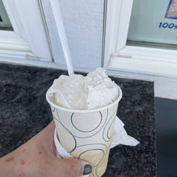 a hand holding a cup of ice cream - A person holds a paper cup filled with vanilla ice cream and a plastic spoon, positioned outdoors near a window with white framing. The hand has painted nails and some small colorful stickers or paint marks. The background includes a black textured surface and part of a white wall and window frame.