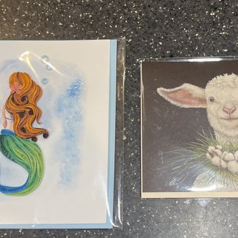 Daily Blooms - Amazing cards towards the back of the store - The image shows two illustrated cards on a dark speckled surface. The left card features a mermaid with long, wavy orange hair, a green tail, and a blue fin, with a background of blue watercolor splashes. The right card depicts a lamb with fluffy white wool, large ears, and a gentle expression, wearing a wreath of white flowers and green grass. Both cards are in clear plastic sleeves. - Home Decor Near Me - Fallbrook, California