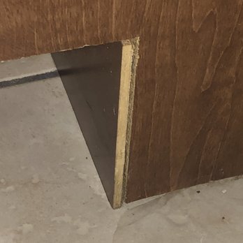 Cole Spring Plaza - bad caulking job it just falls over - The image shows a close-up of a wooden panel with a visible damaged bottom edge, exposing a black triangular support underneath. The panel is positioned on a concrete floor, and the damage appears to be a result of wear or improper installation. - Apartments Near Me - Silver Spring, Maryland
