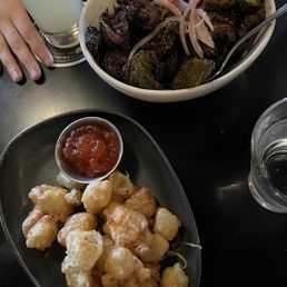 Fried Brussels - The image shows a plate of golden fried cheese curds accompanied by a small cup of red dipping sauce. Next to it, there is a large bowl of roasted Brussels sprouts topped with sliced onions. The setting appears to be a restaurant table with drinks and a person's hand visible.