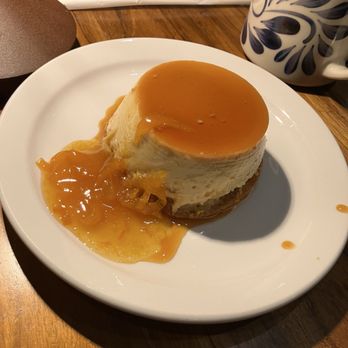 Mercado 925 - Flan - A flan dessert with a smooth, creamy texture is presented on a white plate, topped with a layer of caramel sauce. A portion of caramelized fruit compote is placed beside the flan, with some caramel sauce drizzled on the plate. The setting includes a wooden table, a patterned mug, and part of a gray object in the background. - Mexican Near Me - Berkeley, California