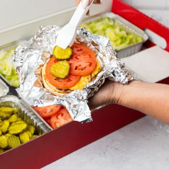 Five Guys - A close-up photo of a person putting pickles on their cheeseburger in front of a Five Guys catering box of toppings. - Burgers Near Me - Mayfield Heights, Ohio Five Guys - A close-up photo of a person putting pickles on their cheeseburger in front of a Five Guys catering box of toppings. - Burgers Near Me - Mayfield Heights, Ohio