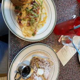 Cinnamon Roll French Toast. - The image shows two plates of breakfast food on a patterned table. The first plate contains an egg white omelet topped with diced ham, green salsa, and melted cheese, accompanied by a baked potato. The second plate holds French toast dusted with powdered sugar, with a small container of syrup on the side. There are also drinks, including a red beverage with a straw and a glass of water, along with sunglasses and a receipt nearby.