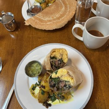 Ama Kitchen - Brunch specials - breakfast burrito and phull dosa. - A meal featuring beef burritos with potatoes and salsa on a white plate, alongside a dosa with chutney, water, coffee, and condiments on a wooden table. - Breakfast & Brunch Near Me - Madison, Wisconsin