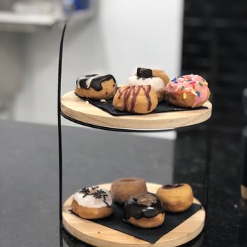 Y&E Creations - a display of donuts - A two-tiered display stand made of wood and black metal holds multiple mini donuts decorated with various toppings and icing. The donuts are arranged on black paper liners on each tier. The background is blurred, focusing attention on the donuts, which include options with chocolate, pink icing, sprinkles, and other toppings. The setting appears to be inside a shop or cafe. - Desserts Near Me - Middletown, New York