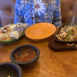 Fajita - A person wearing a plaid shirt is seated at a wooden table in a casual Mexican restaurant. The table has a plate of fajitas with chicken and vegetables on a sizzling skillet, a bowl of rice with vegetables and a foil-wrapped item, and a small bowl of salsa. An empty orange plate is also on the table. The background shows a booth with padded seating and a handbag hanging on the side.