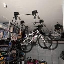 Bike Pulleys - A white mountain bike is hanging from a ceiling-mounted bike hoist in a garage, with storage shelves and various toys and tools visible.