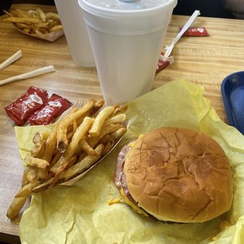 Storm's - Lunch. - The image shows a cheeseburger with a toasted bun, melted cheese, and a beef patty on yellow paper. Next to it is a paper tray filled with French fries. Two large white cups with lids and straws are also on the table, along with some ketchup packets and utensils. The setting appears to be a casual dining restaurant with a wooden table surface. - Fast Food Near Me - Hamilton, Texas