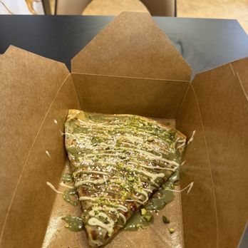 Cafe Bazaar - Dubai Crepes - A pizza slice topped with green sauce, white drizzle, and crushed pistachios, served in a brown takeout box on a black table. - Cafes Near Me - Brooklyn, New York