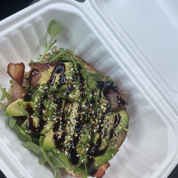 CoffeeHead - Avocado Toast w/bacon on Sourdough - A piece of toast topped with avocado, bacon, arugula, sesame seeds, and drizzled with balsamic glaze, served in a white takeout container. - Coffee & Tea Near Me - Topeka, Kansas