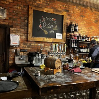 Debajo Tapas ABQ - a man at a bar - The image shows the interior of a tapas restaurant with a brick wall and a wooden-framed chalkboard displaying drawings of food and drinks. Shelves filled with wine bottles are visible on the right side. A wooden bar counter with a small barrel, glasses, and decorative plants is in the foreground. A staff member is working behind the bar, and a sink with glasses is also visible. The setting suggests a casual dining atmosphere focused on Spanish-inspired cuisine. - Spanish Near Me - Albuquerque, New Mexico