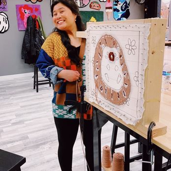 Tuft A Rug - All handmade rugs on the wall behind me! - A woman with long dark hair, wearing a colorful patchwork sweater, stands beside a tufted rug artwork in a craft studio with colorful wall decorations. - Art Classes Near Me - Aurora, Illinois