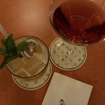Double Fun - The image depicts two drinks on a table: a cocktail with ice, mint leaves, and lemon slices, and a glass of red wine. The drinks are placed on coasters, and a paper with the words 'Double Fun' and a cherry illustration is also visible. The table surface is a reddish-brown color. - Cocktail Bars Near Me - Chicago, Illinois