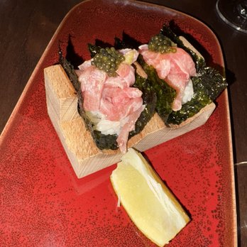 Blu Ember - Toro Nori Bites - Two pieces of sushi are presented on a red plate, each topped with slices of raw fish, seaweed, and green caviar. The sushi is placed on a small wooden block, and a lemon wedge is positioned nearby. The setting appears to be a dining table at a Japanese restaurant, with a dark background and a glass of wine partially visible. - New American Near Me - Queens, New York