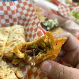 Birria Ramen Soup with 1 Quesabirria Taco - A person holds a quesabirria taco filled with meat, vegetables, and cheese, with other food items and red checkered paper in the background.