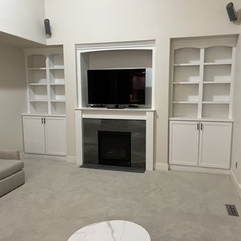 J Brothers Construction - Living room featuring a fireplace with a mounted TV above, flanked by built-in white shelves and cabinets, with a beige sofa on the left. - General Contractors Near Me - Gresham , Oregon