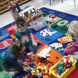 Different learning centers for everyone! - Three children are seated on a colorful educational rug in a classroom, engaging with various toys including building blocks, stuffed animals, and toy vehicles. The rug features alphabet letters and images, and there are shelves and chairs in the background, indicating a preschool or early childhood learning environment.