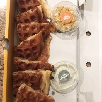 Waffadilla - Classic waffadilla - A row of waffle fries is arranged on a white tray, accompanied by two small containers of dipping sauces labeled 'Corn Relish' and 'Spinach Artichoke.' The fries are golden brown and crispy, with some spilling slightly onto the tray. The sauces are in clear plastic containers, with the labels indicating their flavors. The tray is placed on a brown surface, and part of a white box is visible in the background. - Waffles Near Me