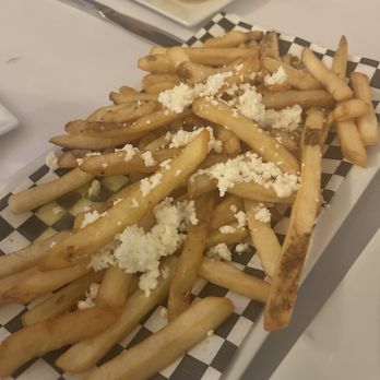 Pete's Taverna - Fries, lemon oil and feta. OMG!!! - French fries topped with crumbled feta cheese are presented on a checkered paper-lined tray, with a small dish of lemon oil and a potato wedge in the background. - Cocktail Bars Near Me - Riverhead, New York