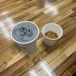 midnight rose - Two paper cups of coffee sit on a wooden table, one with foam and black seeds, the other with espresso remnants.