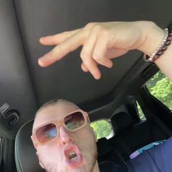 Seven Days - Party in my mf tummy - A young man with light skin, short hair, and a beard is seated in the backseat of a car, wearing large, pink-tinted sunglasses. He is making a playful, puckered-lip expression and holding up his right hand in a peace sign gesture. The car interior features a black headrest and a window showing green foliage outside. The man is wearing a dark shirt and has a bracelet on his right wrist. - Breakfast & Brunch Near Me - Dennis, Massachusetts