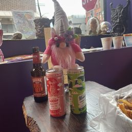 Our Travel Gnome Skye enjoying a refreshing Saranac Root Beer - A plush unicorn gnome toy with a flower crown and a horn is on a table alongside cannabis-infused drinks and a beer bottle, with a window showing street scenery.